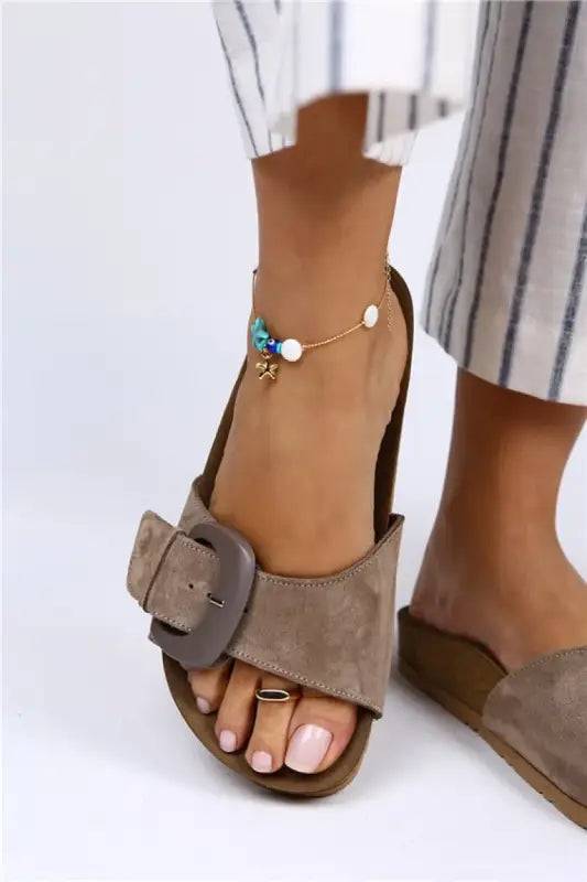 Gray suede sandal with a prominent square buckle and an ankle bracelet visible on the wearer’s foot.