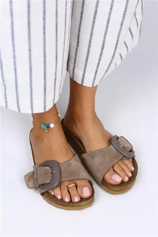 Taupe suede sandals with decorative buckles and a flat sole.