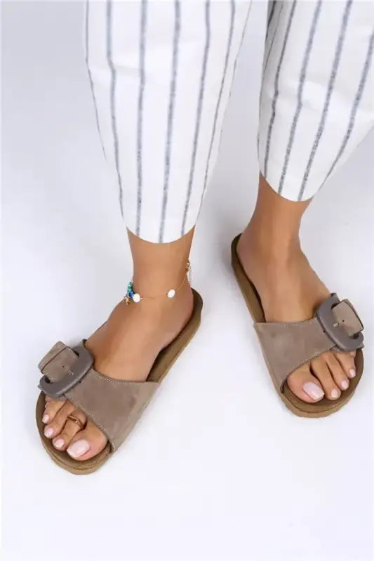 Taupe suede sandals with decorative buckles and a flat sole.